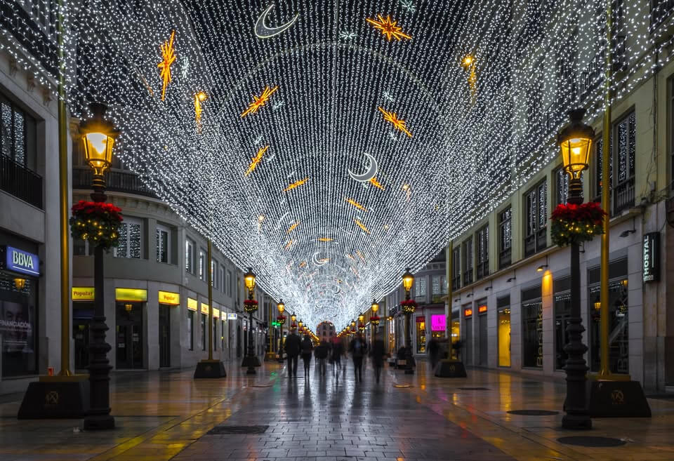 A festive nighttime street scene with a tunnel of white string lights and glowing decorative lights overhead, lined with illuminated lampposts and storefronts
