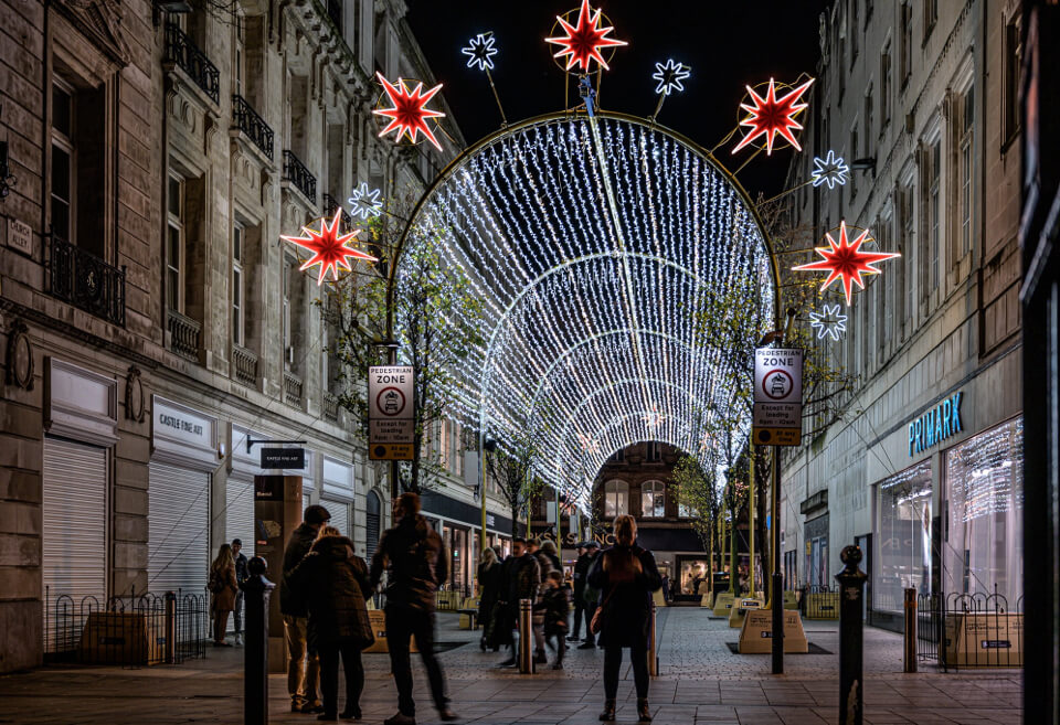 A nighttime street scene under a large arched tunnel of white holiday lights, with red star-shaped decorations hanging overhead and pedestrians walking along between buildings